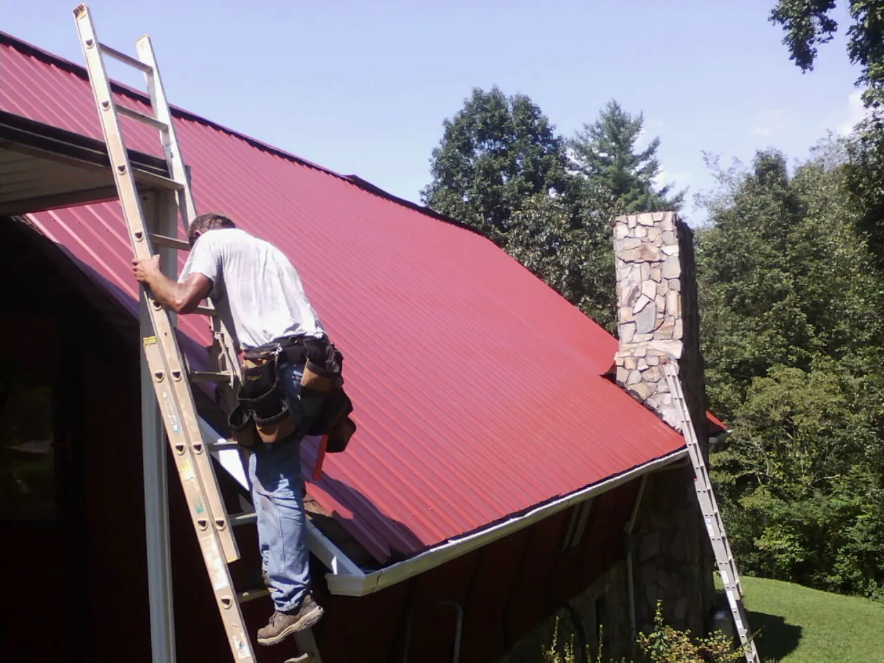 Roofer on a ladder beside a red metal roof and stone chimney — Soffit & Fascia Repair by Roofing Brothers in Marriottsville